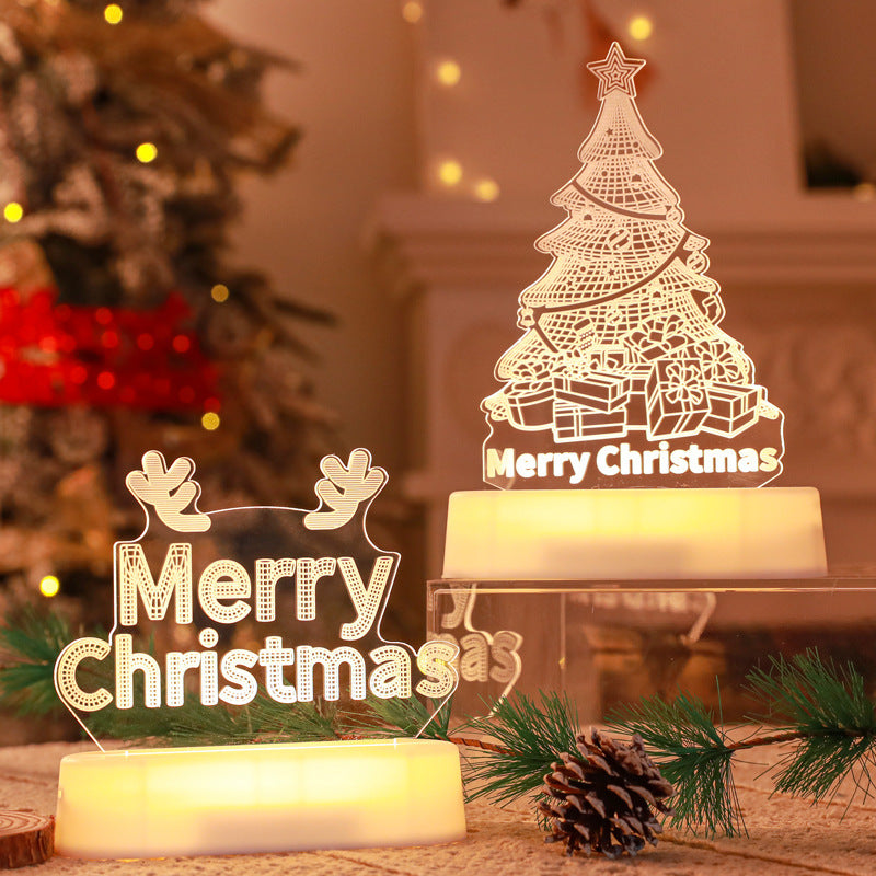 Illuminated Christmas LED lamps with a 'Merry Christmas' sign featuring reindeer antlers and a Christmas tree with presents underneath. The lamps glow warmly, set against a backdrop of a decorated Christmas tree with lights and ornaments. Pine branches and a pine cone add to the festive ambiance.