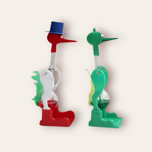 Original Drinking Bird toy, featuring two plastic birds with felt-covered heads and midsections that resemble feathers. The bird on the left is red with a blue hat and white ‘feathers’, while the bird on the right is green with a yellow beak and yellow ‘feathers’. Both birds have clear glass tubes extending from their tails to their beaks, filled with a colored liquid. Ideal for Giggly Groves’ novelty toy collection.